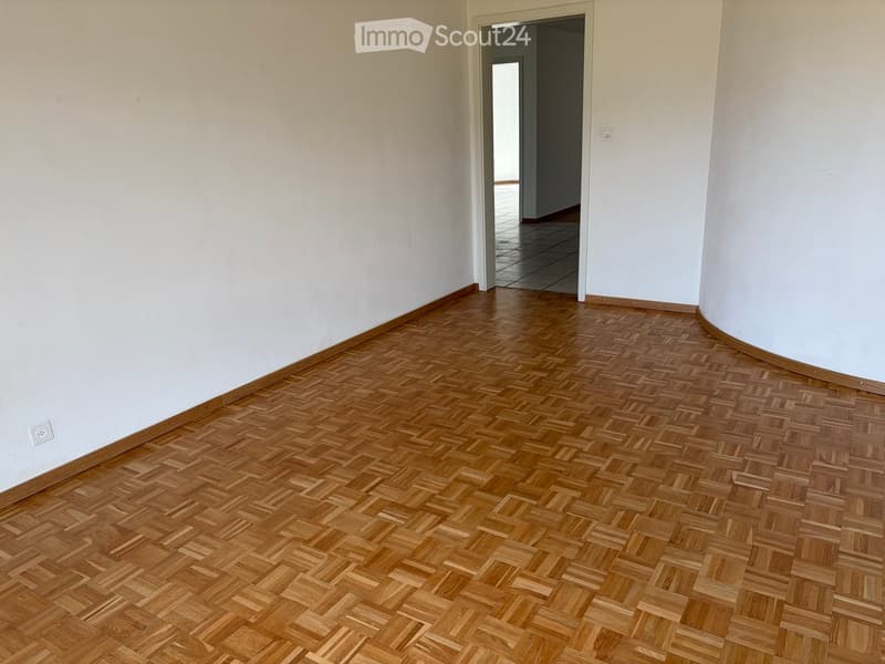 Empty hallway with wooden herringbone flooring and white walls, featuring an open doorway leading to another room.