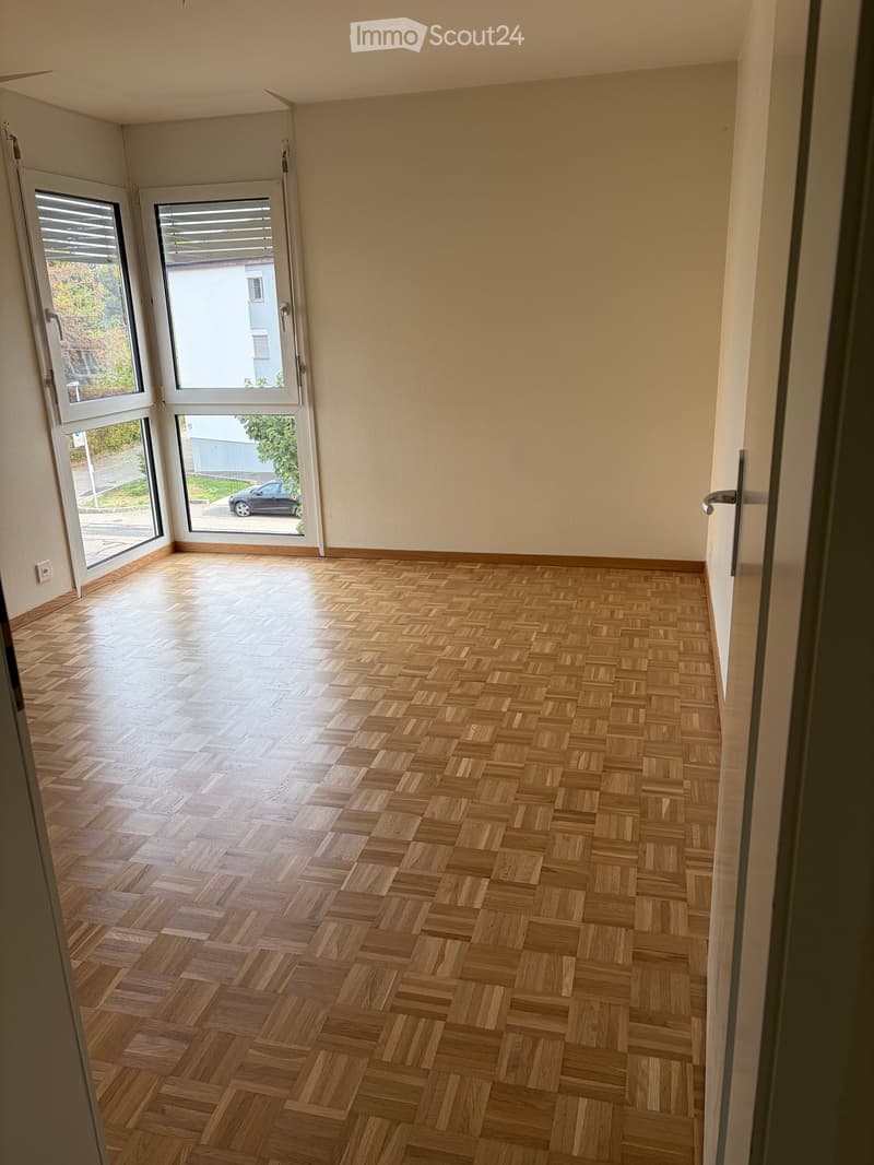 Empty room with wooden herringbone flooring, large windows with blinds, and white walls.