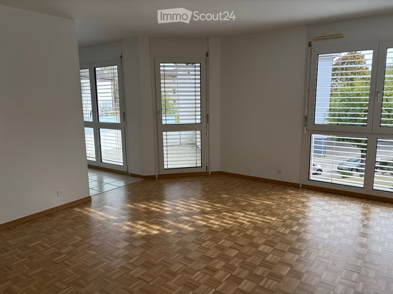 Empty room with wooden parquet flooring, large windows with blinds, and white walls.