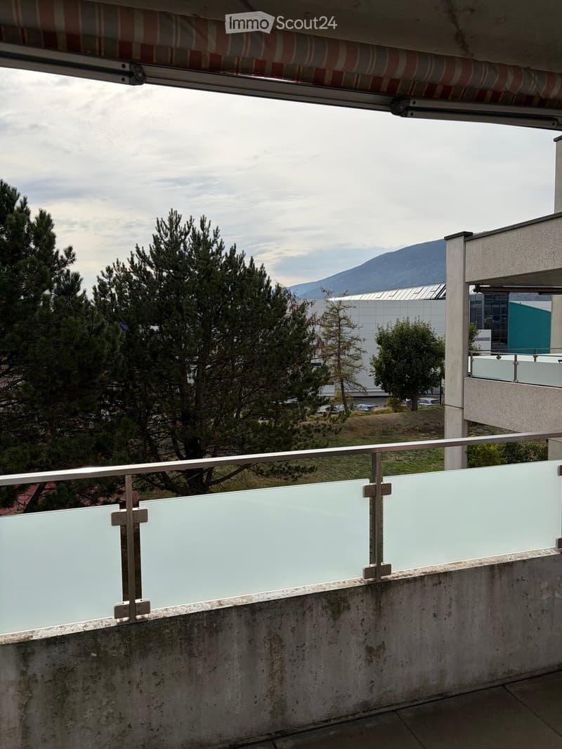 Balcony with glass railing, overlooking a landscaped area with trees and a distant mountain view.