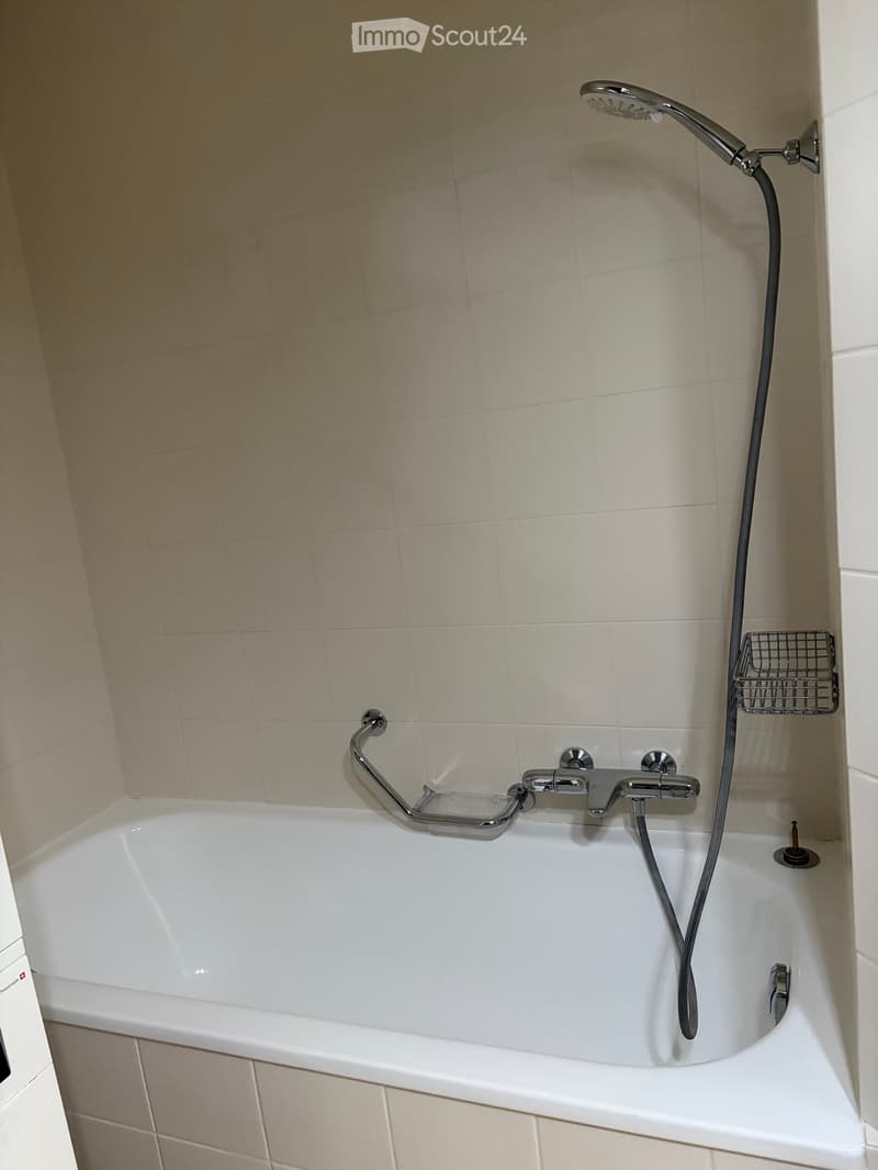 Bathtub with a handheld showerhead and a built-in handrail, surrounded by beige tiled walls.