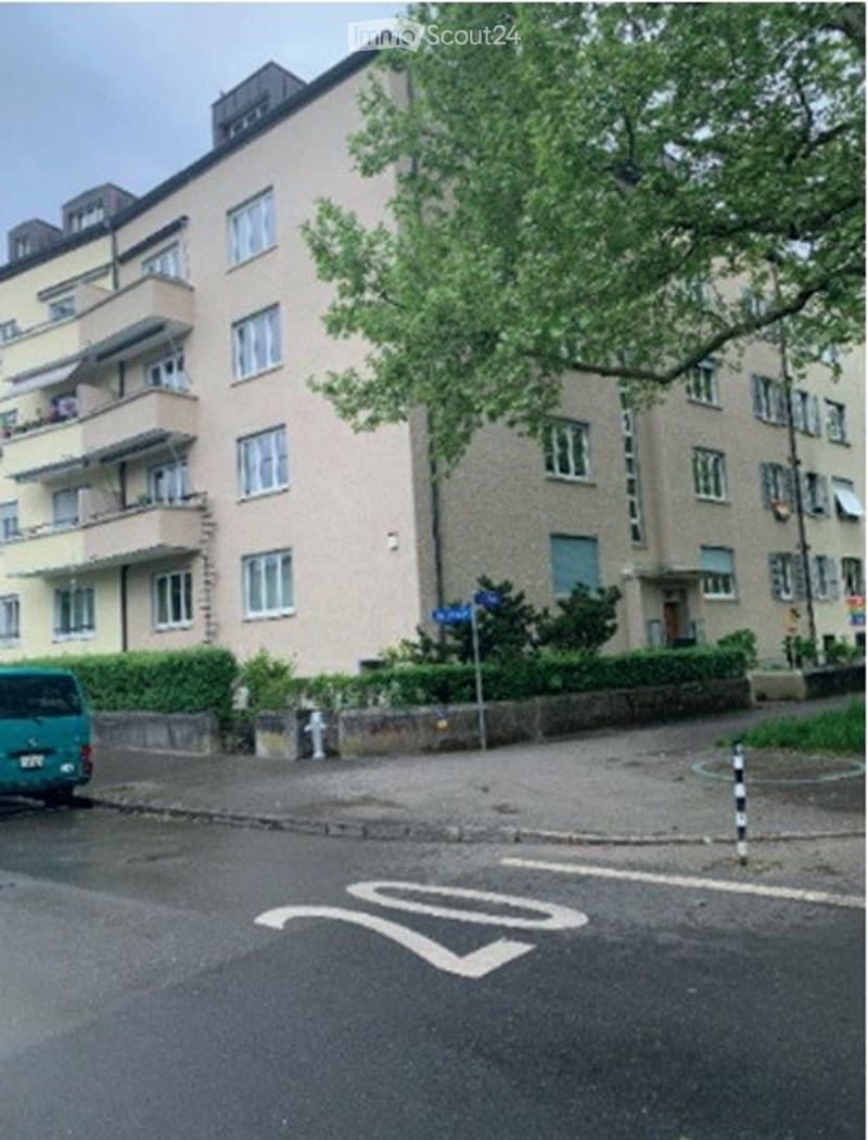 Multi-story residential building with multiple windows and balconies, surrounded by greenery and a parked van.