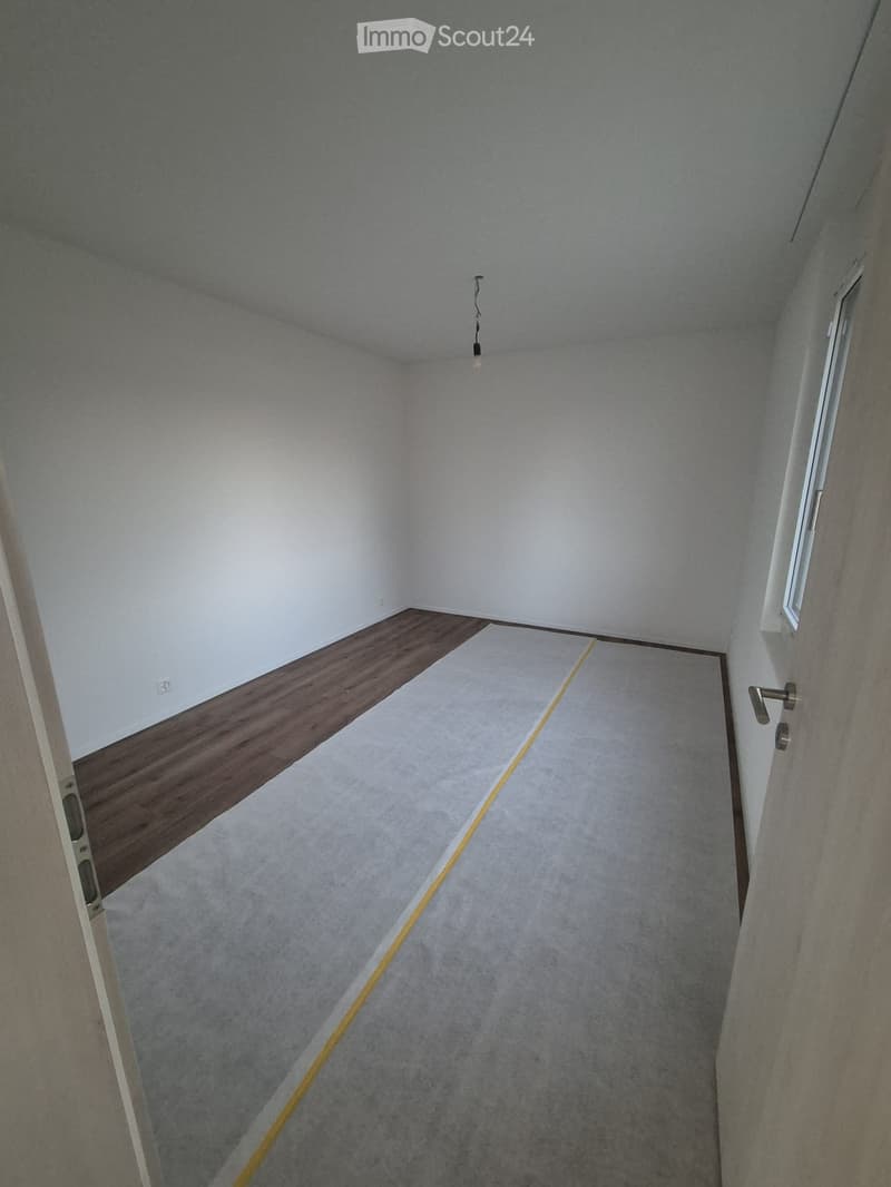 Empty room with white walls, grey carpet, wooden shelf, and a single light fixture hanging from the ceiling.