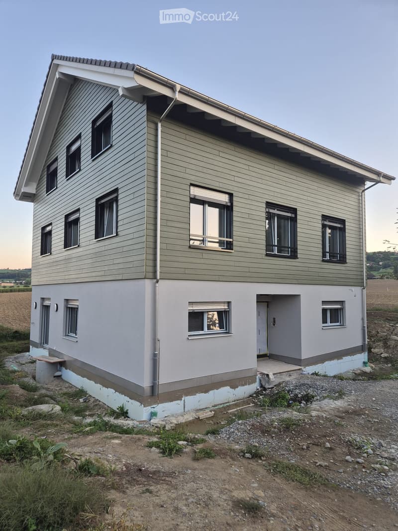 Two-story house with green siding, multiple windows, and a raised ground floor. The house features a modern design with a sloped roof and a small porch area.