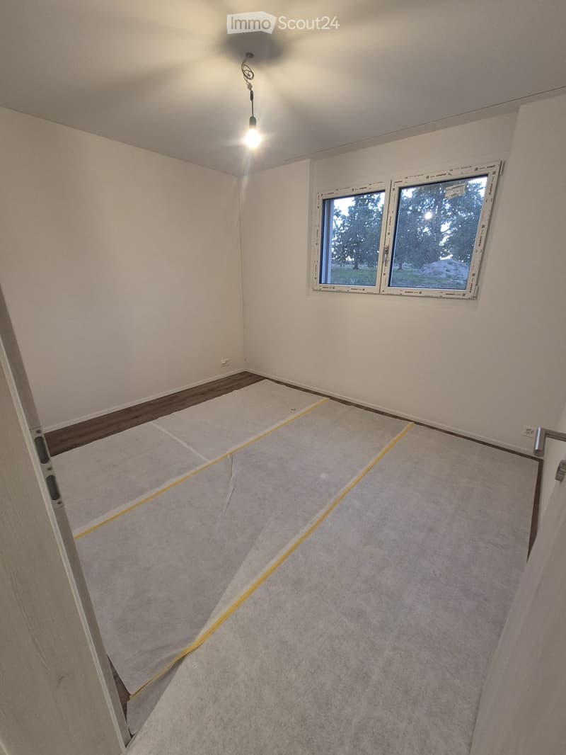 Empty room with bare walls, wooden baseboard, and a single window with white frame. The floor is covered with a protective sheet and yellow tape, indicating preparation for renovation.