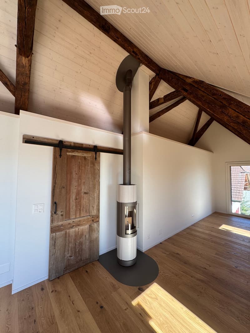 An empty attic space with exposed wooden beams, a modern wood-burning stove, and a large wooden sliding door.