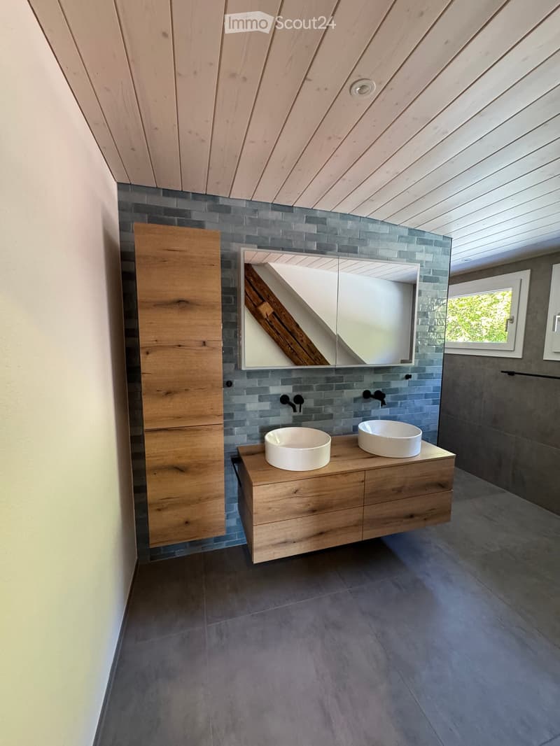 Modern bathroom with wooden vanity, two sinks, grey tiled walls, and a large mirror.