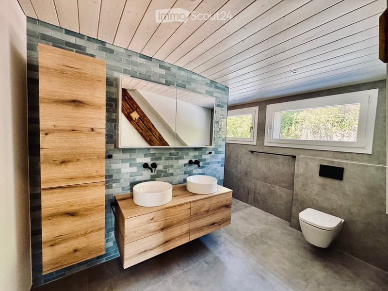 Modern bathroom with wooden vanity, two sinks, a large mirror, and a toilet. The walls are tiled in a mix of grey and blue tiles, and the ceiling has a wooden slatted design.