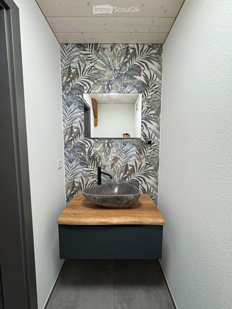 Modern bathroom with a wooden countertop, stone sink, black faucet, and a mirrored cabinet above. The walls feature a leafy, tropical print design.