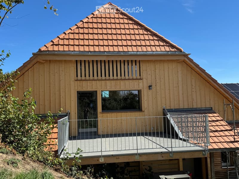 Modern wooden house with a raised ground floor, large windows, and a balcony with metal railing.