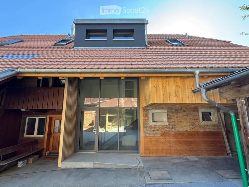 Two-story house with a tiled roof, wooden siding, and a modern glass entrance. Features include a raised ground floor, a small wooden bench, and a brick wall section.