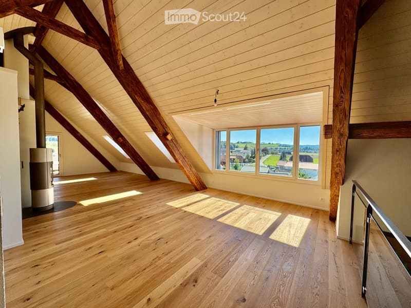 Bright, spacious attic room with wooden floors, exposed wooden beams, large windows offering a scenic view, and a modern wood-burning stove.
