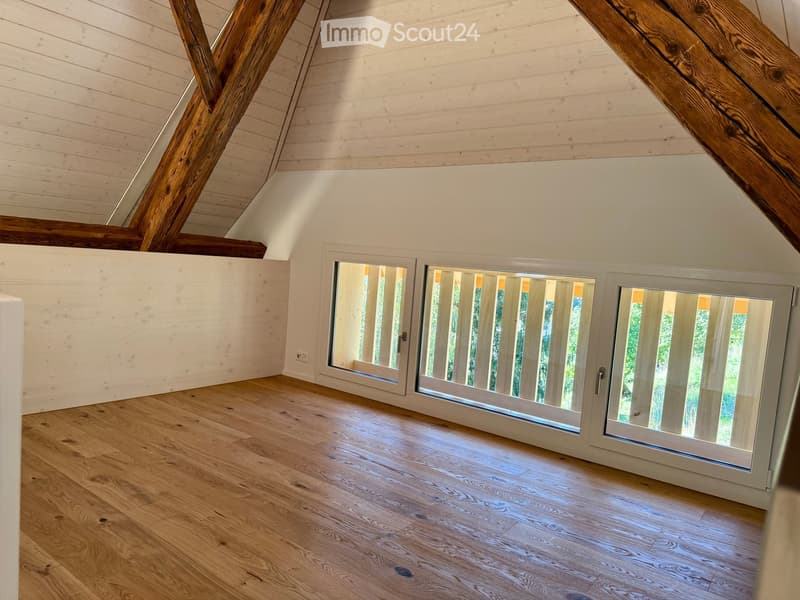 An empty attic space with wooden beams, white walls, and large windows offering a view of greenery outside. The floor is made of light wooden planks.