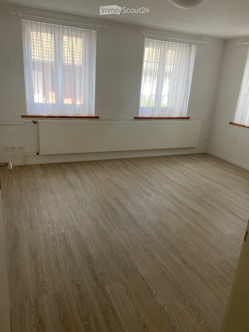 Empty room with wooden flooring, white walls, and large windows with white curtains. A radiator is mounted on the wall below the windows.