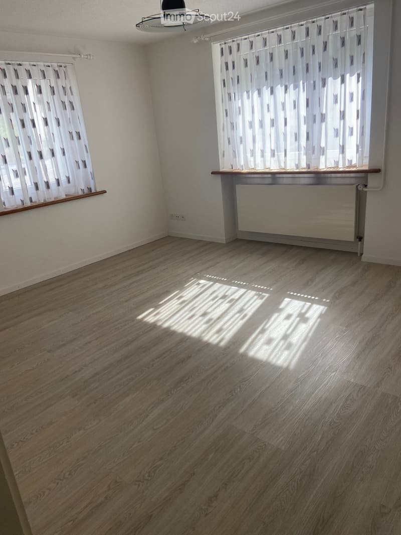 Empty room with wooden flooring, white walls, and two windows with white curtains. A radiator is visible under one window, and a ceiling fan is mounted on the ceiling.