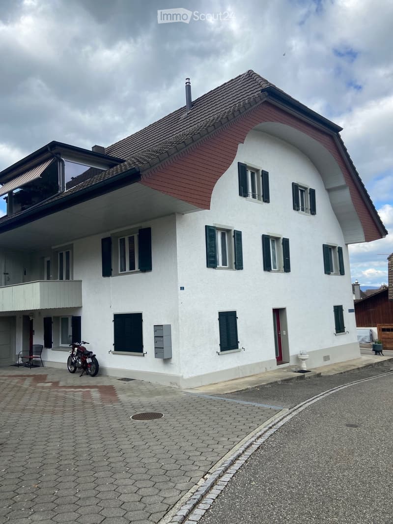 Two-story house with white walls, red roof, multiple windows with green shutters, a balcony on the upper floor, and a motorcycle parked in front.
