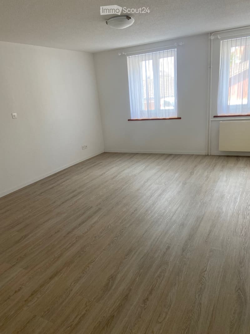 Empty room with wooden flooring, white walls, and large windows with white curtains. There is a radiator on one wall and a light fixture on the ceiling.