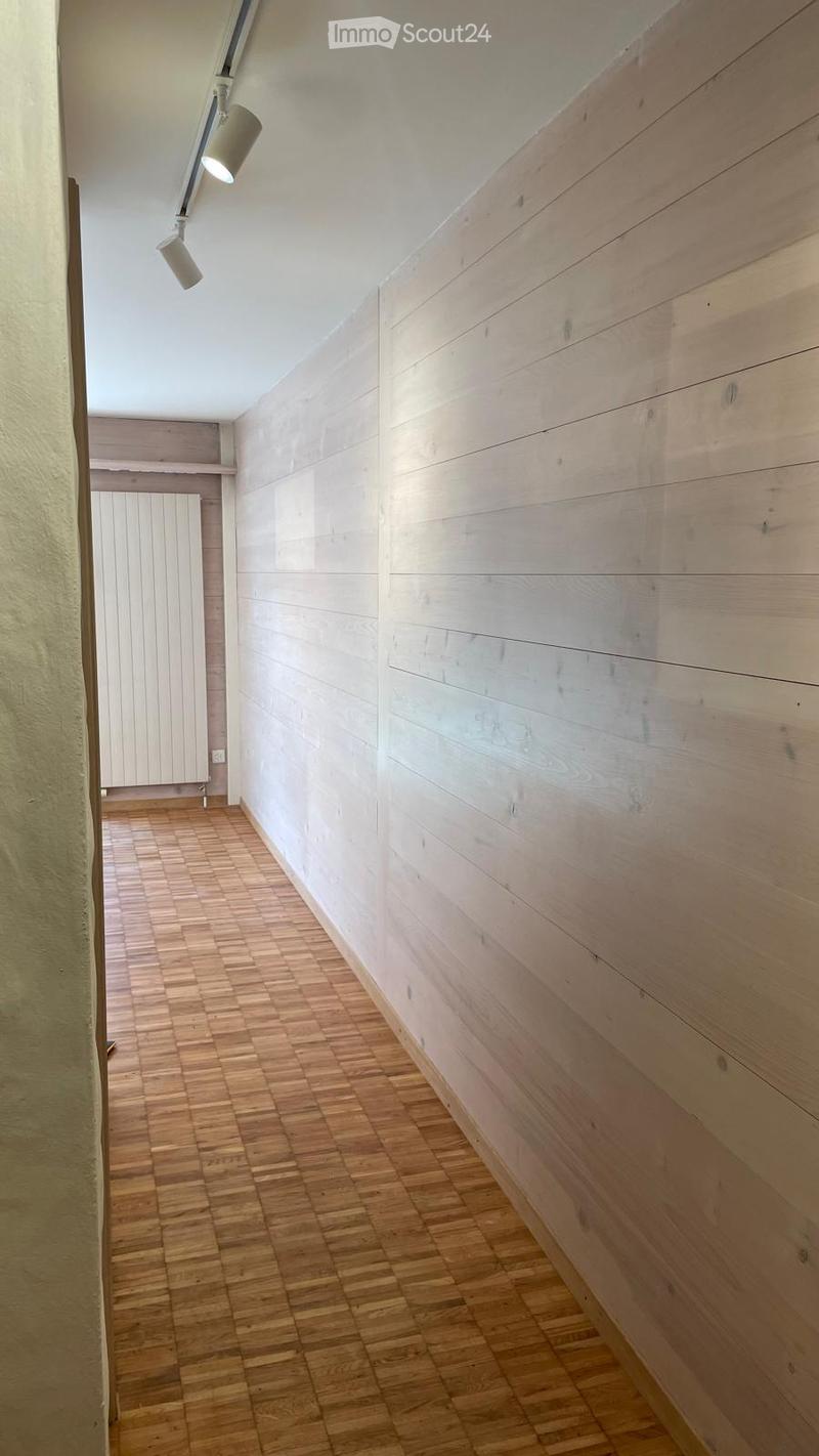 A narrow hallway with wooden panel walls and a herringbone-patterned wooden floor, illuminated by a wall-mounted light fixture.