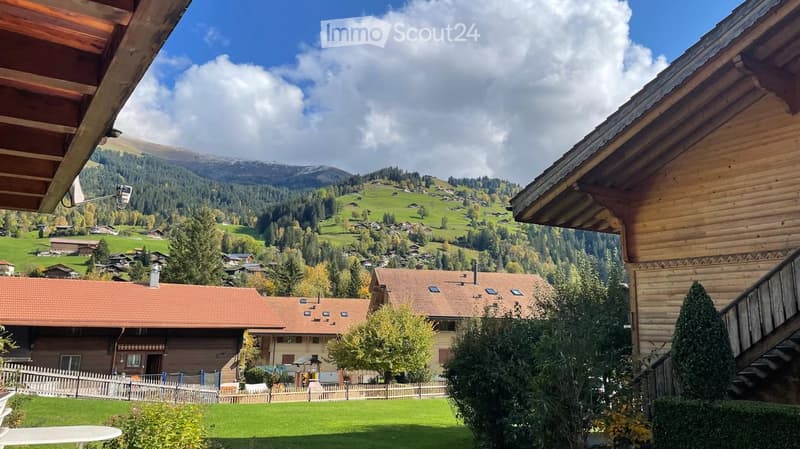 A scenic outdoor view from a wooden balcony overlooking a picturesque village with traditional houses, green fields, and distant snow-capped mountains.