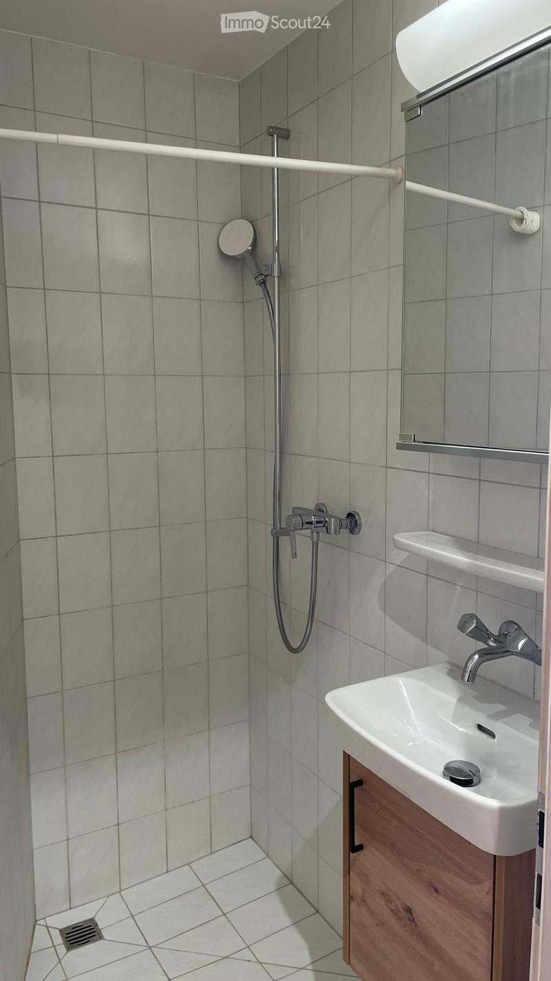 Modern bathroom with grey tiled walls and floor, a white sink with a wooden cabinet, a showerhead with a rail, and a mirror above the sink.