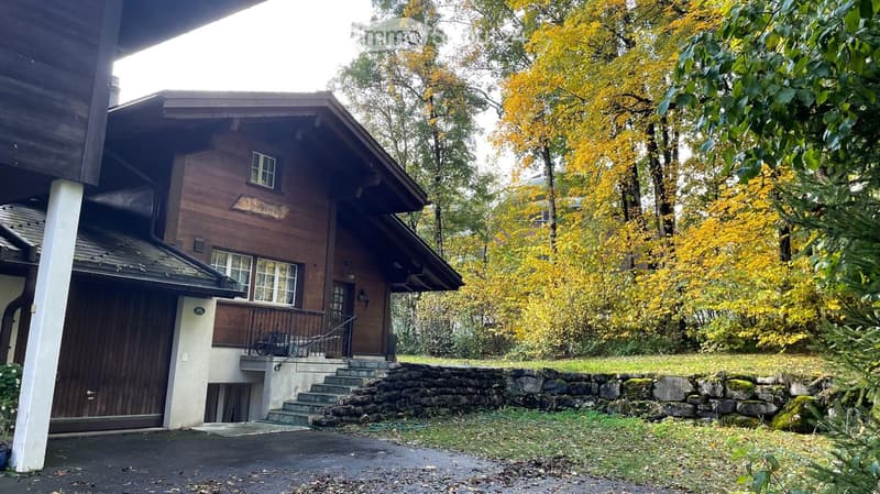 A two-story wooden house with a stone wall and steps leading to the entrance, surrounded by autumn foliage and trees.