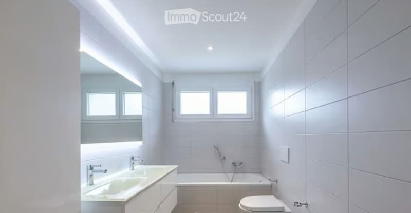 Modern, minimalist bathroom with white tiles, two sinks, a bathtub, and a toilet. Features large windows and sleek lighting.