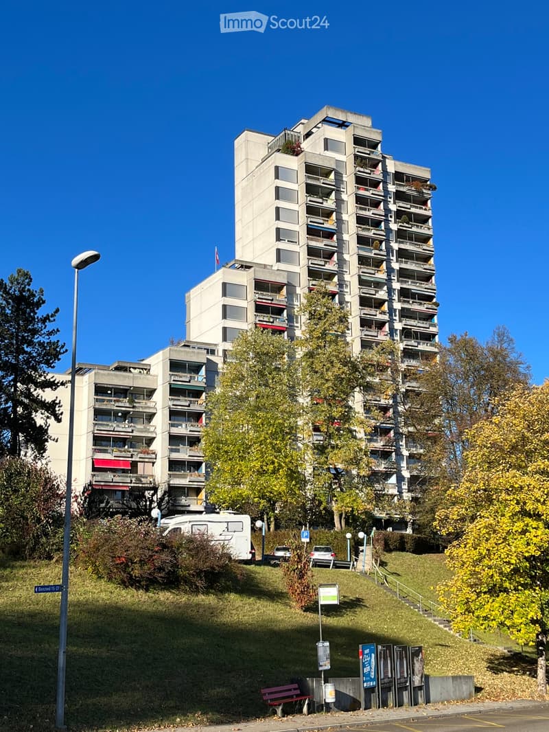 Multi-story residential building with balconies, surrounded by trees and a grassy slope with a bench and parked vehicles.