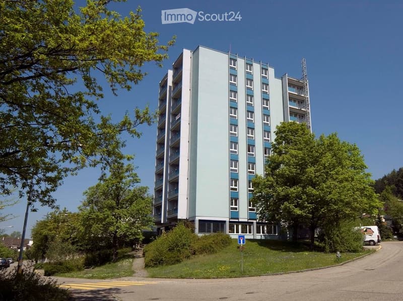 A modern multi-story apartment building with light blue and white facade, numerous windows, and balconies. Surrounded by greenery and trees.