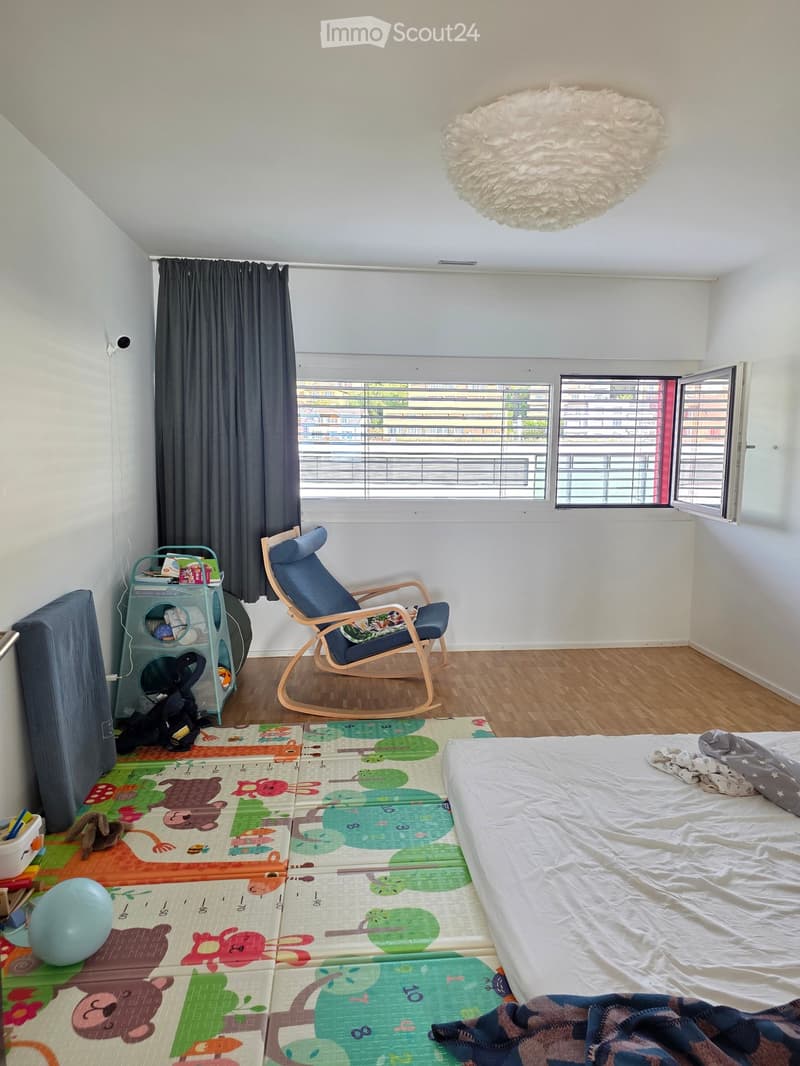 Bright bedroom with wooden flooring, large window with open blinds, bed with white sheets, colorful play mat, rocking chair, storage unit, and decorative lamp.