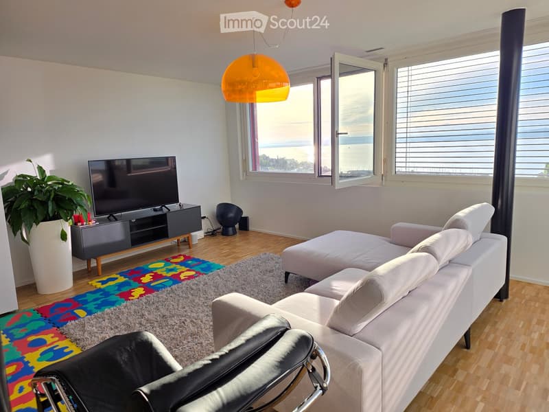 Modern living room with a white sofa, black leather chair, TV on a stand, colorful rug, and a large window offering a scenic view.