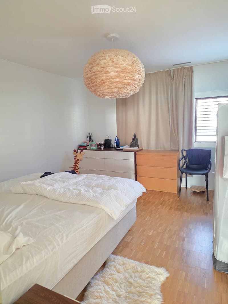 A well-lit bedroom with a large bed, a wooden floor, a fluffy white rug, a hanging round lamp, a small table with various items, a blue chair, and a window with curtains.