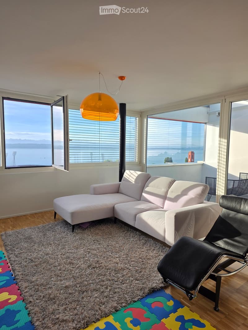 Modern living room with a white sectional sofa, black leather chair, large windows offering a scenic view, and a colorful rug on the wooden floor.