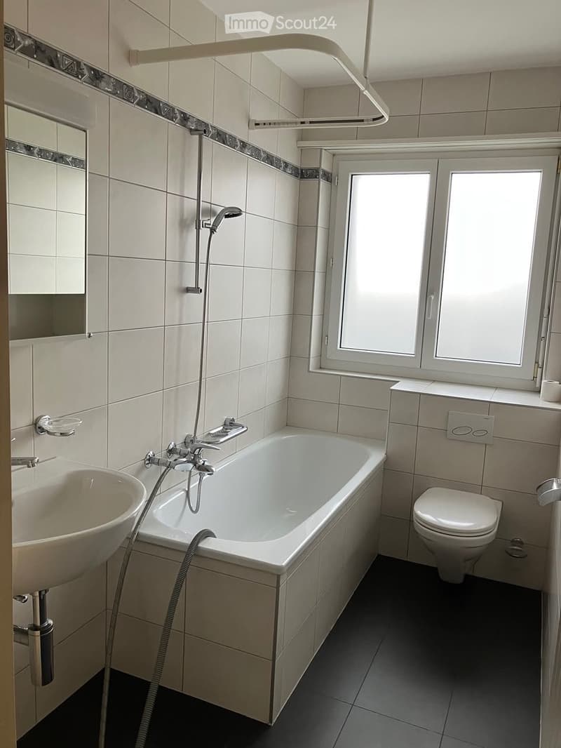 Modern bathroom with a bathtub, toilet, sink, and showerhead. The room features tiled walls and a window providing natural light.