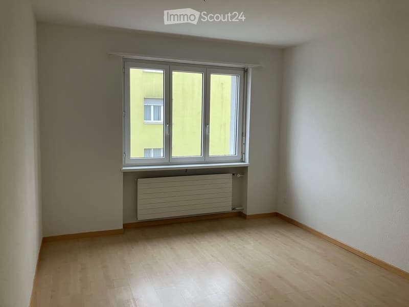 Empty room with wooden flooring, white walls, a large window with white frame, and a radiator below the window.