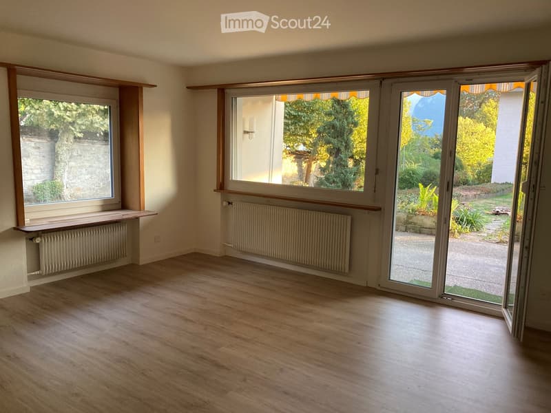 Empty room with wooden flooring, large windows, radiators, and sliding glass doors opening to a garden.