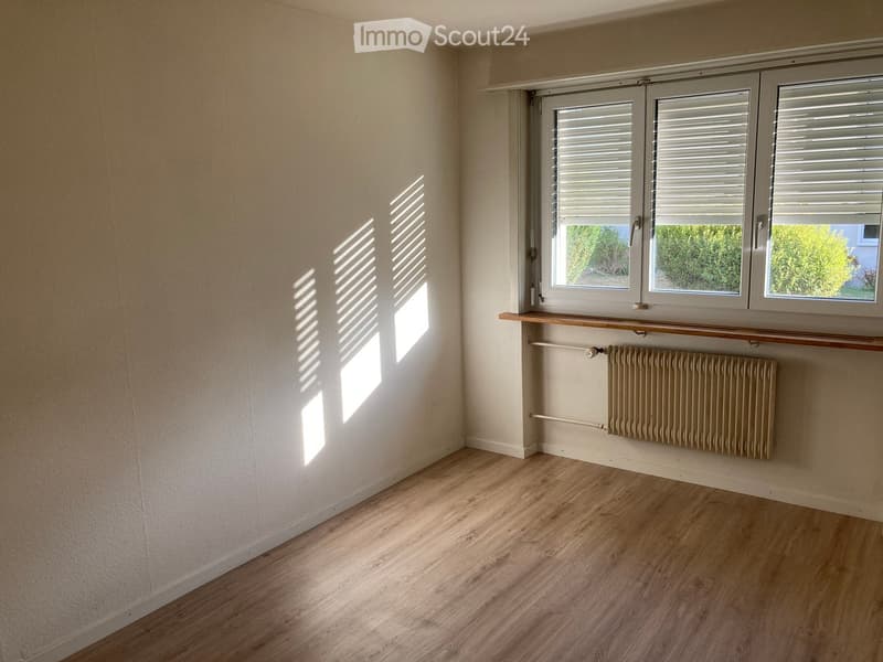 Empty room with light wooden flooring, white walls, and a window with blinds. A radiator is visible below the window, and a small wooden shelf is mounted on the wall above it.