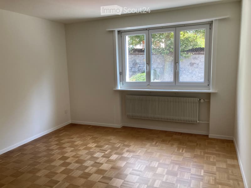 Empty room with wooden flooring, white walls, and a large window with a radiator below it.