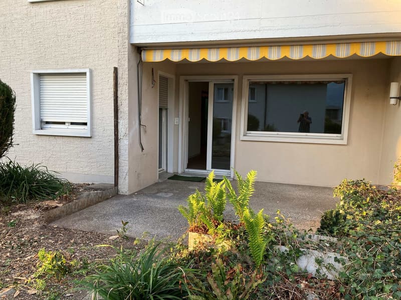 Two-story building with a covered entrance, windows, and small garden area with plants and shrubs.