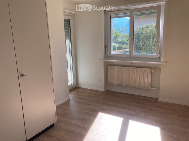 Empty room with light wooden flooring, white walls, and large windows offering a view of greenery. A radiator is mounted below one window, and a door is visible on the left side.