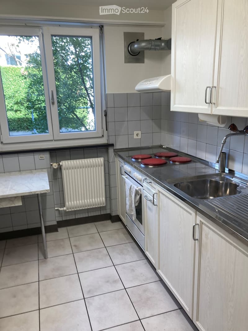 Modern kitchen with white cabinets, tiled walls, and a stainless steel sink. Features an electric stovetop, a radiator, and a small table with a marble top.