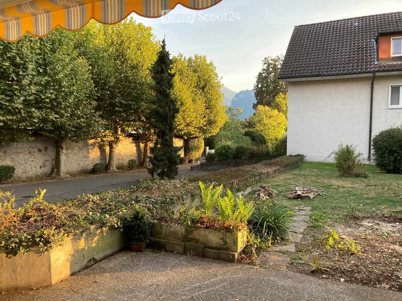 Garden area with gravel pathway, raised planter boxes, trees, and a stone wall. A house with white walls and a dark roof is visible in the background, along with a view of mountains.