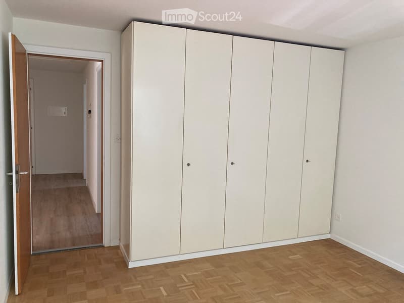 Empty room with light wooden flooring, white walls, and a large built-in white wardrobe unit. An open doorway leads to a hallway.