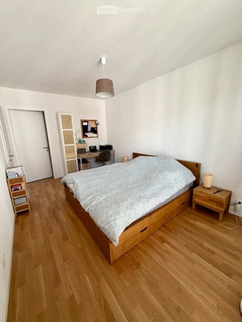 Cozy bedroom with wooden flooring, a large bed with a grey duvet, a nightstand with a lamp, a desk with a chair, and a shelf with books.
