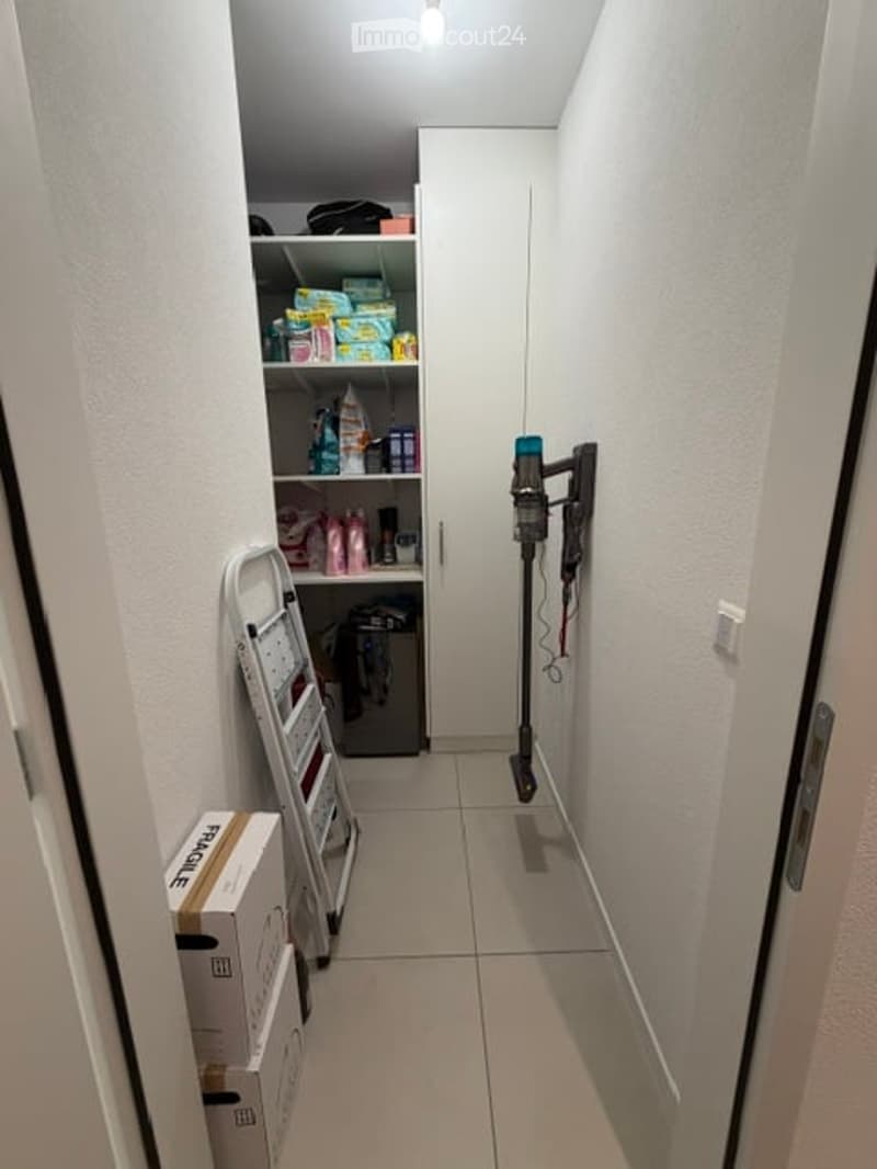 Narrow hallway with white walls, tiled floor, and a small built-in shelf unit containing various items. A vacuum cleaner is mounted on the wall, and a ladder and boxes are placed on the floor.