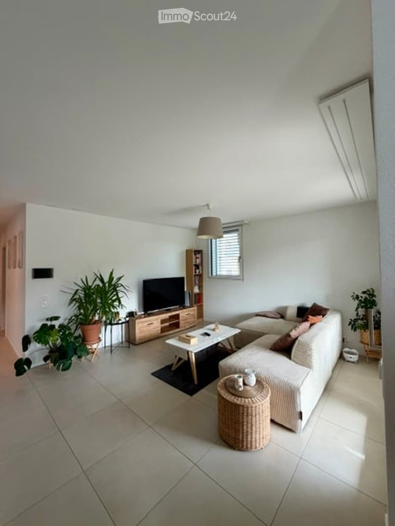 Bright living room with white walls, tiled floor, and large windows. Features a cream sectional sofa, coffee table, TV on a wooden stand, side tables, and potted plants.