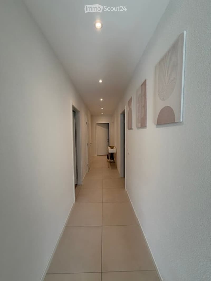 Clean, well-lit hallway with white walls, beige tiled floor, and modern artwork on one side.