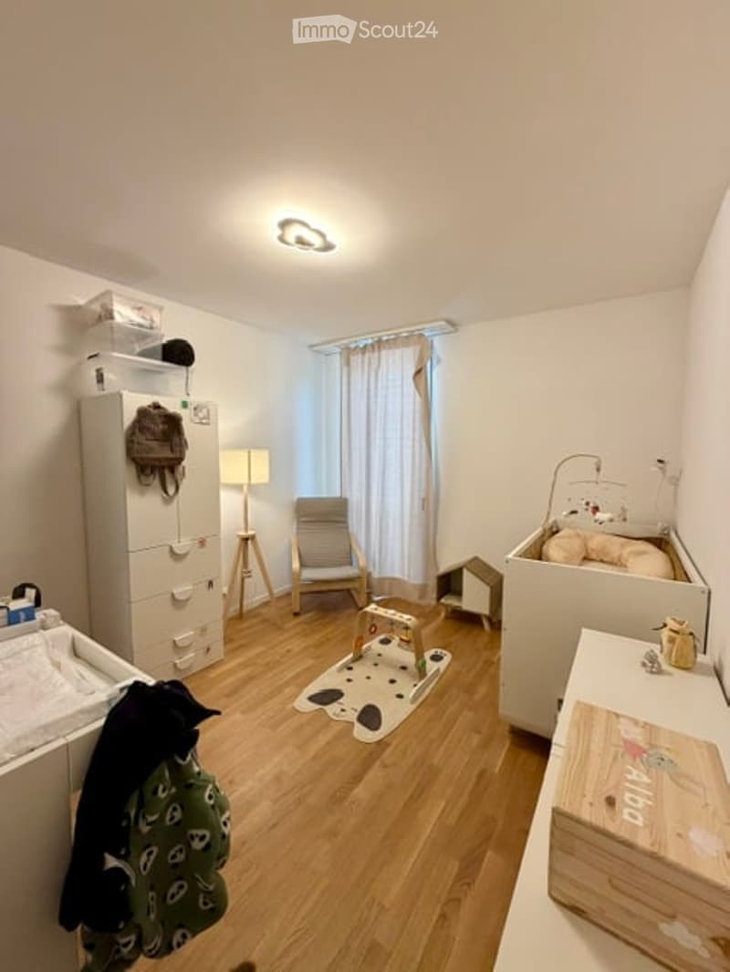 A well-lit nursery with wooden flooring, a crib, a changing table, a small chair, a lamp, and a cabinet. The room features a curtained window and a small play area with a toy and a stool.