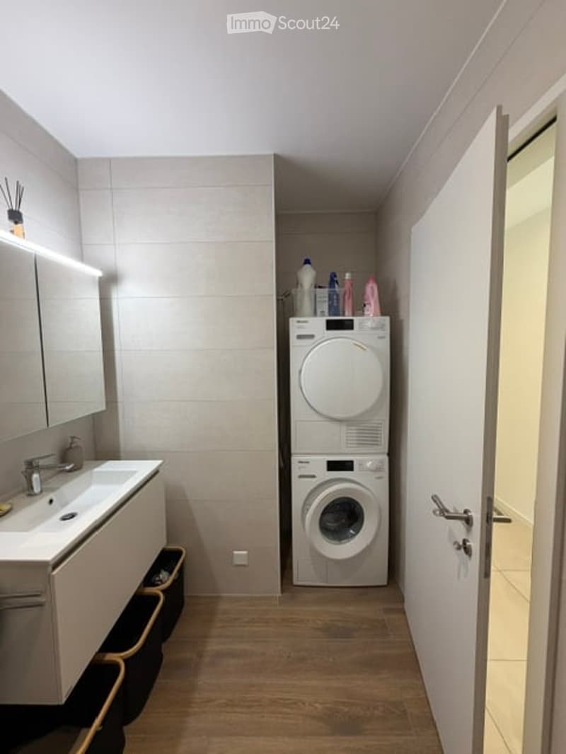 Modern bathroom with a sink, mirror, and a stacked washer/dryer unit. The space features light-colored tiles and a wooden floor.