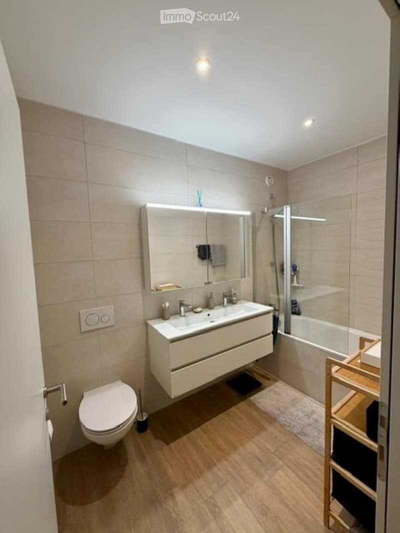 Modern bathroom with a white sink, large mirror, toilet, and bathtub. Features include a wooden floor, beige tiled walls, and recessed lighting.