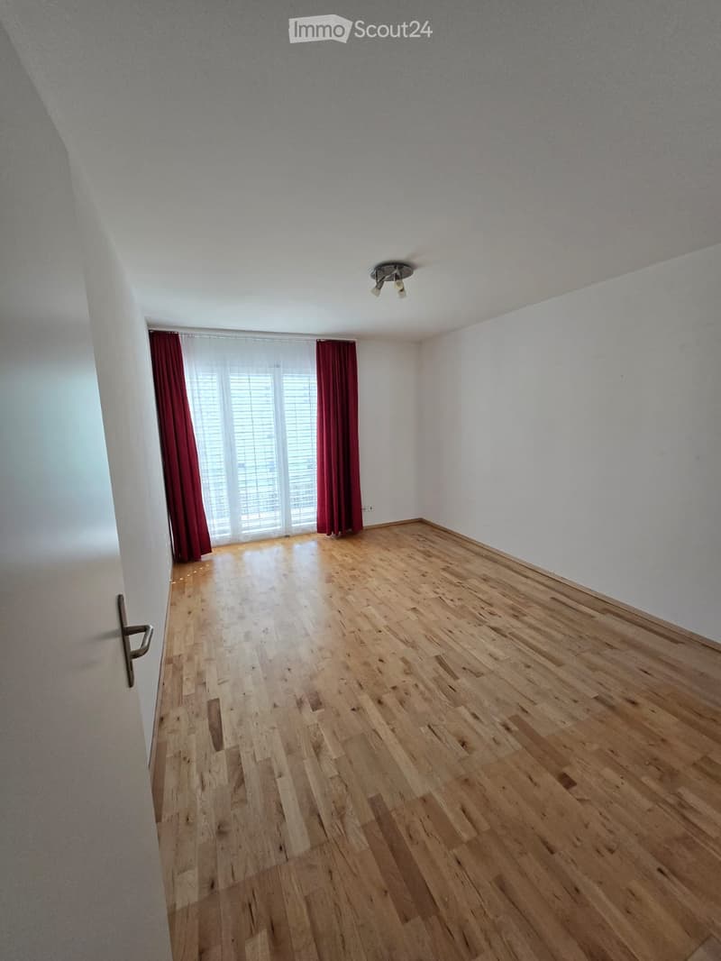 Empty room with wooden flooring, white walls, and large windows with red curtains. A door is visible on the left side.